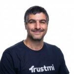 A man with short dark hair and a beard smiles at the camera, wearing a navy blue Trustmi t-shirt, confidently showcasing the brand against a plain white background.