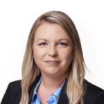 A woman with long blonde hair, wearing a black blazer and a blue shirt, smiles slightly at the camera against a plain white background, conveying the professionalism and confidence expected during bank account verification.